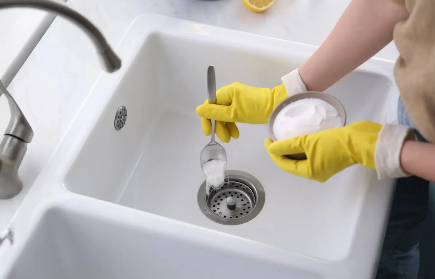 Personne portant des gants jaunes versant du bicarbonate de soude dans un évier pour déboucher ou entretenir les canalisations.