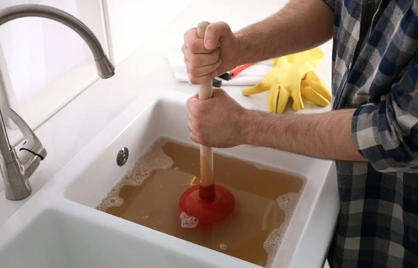 Personne utilisant une ventouse pour déboucher un évier rempli d'eau stagnante, avec des gants et des outils de plomberie en arrière-plan.